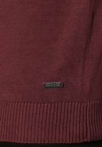 Bourgondische gebreide trui met geribde zoom en een metalen label met daarop "EDC & CO" dicht bij de onderkant. Soepele textuur en nauwsluitend ontwerp.