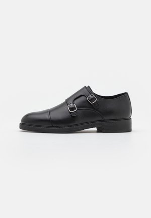 Chaussure habillée en cuir noir avec deux boucles métalliques, bout arrondi, texture lisse et semelle en caoutchouc à profil bas.
