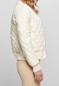 Chaqueta bomber de crema acolchada con puños y dobladillo acanalados, que presenta una cremallera frontal y bolsillos laterales, destacando una textura acolchada y un patrón de diamantes.