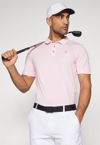 Rosa pikétröja med krage, treknapps knäppning och CK-logotyp, tillsammans med vita byxor och ett svart bälte, medan han håller en golfklubba.
