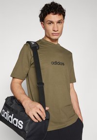 Olivově zelené bavlněné tričko s krátkým rukávem a černým logem adidas na hrudi; v kombinaci s černým sportovním tahem s bílým logem.