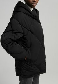 Czarna pikowana parka z dużym kapturem, skośnym pikowaniem i bocznymi kieszeniami. Materiał wydaje się miękki i ocieplany. Projekt o długości do połowy uda.