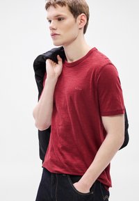 Κόκκινο t-shirt με στρογγυλή λαιμόκοψη από ελαφρύ ύφασμα με διακριτή υφή, που διαθέτει ένα μικρό κεντητό λογότυπο στο στήθος. Σκούρο τζιν και μαύρο μπουφάν.