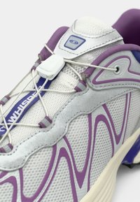 Gray athletic shoe with purple accents, featuring mesh material, unique wave-like design, elastic laces, and a white lacing lock.