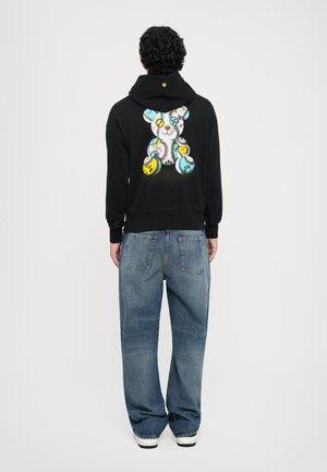 Uomo con cappuccio nero e una grafica di un orsetto colorato ricamato sulla schiena, jeans blu e sneaker bianche, in piedi di spalle.