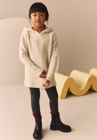 Pull en tricot crème à capuche au toucher doux, associé à un legging noir et des bottines noires. L'arrière-plan présente un design ondulé jaune.