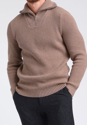 Mann trägt einen beige gerippten Strickpullover mit einem Viertel-Reißverschluss-Kragen, kombiniert mit dunklen, strukturierten Hosen, Hände in den Taschen.