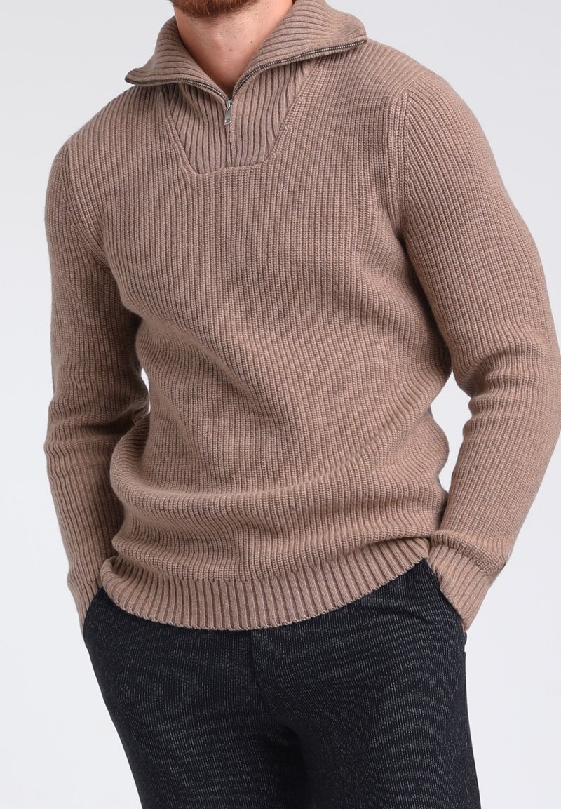 Homme portant un pull en maille côtelée beige avec un col à fermeture éclair quart, associé à un pantalon texturé foncé, les mains dans les poches.