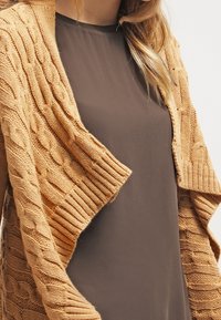 Sweter rozpinany z dzianiny w ciepłym, jasnobrązowym kolorze z otwartym przodem, noszony na gładkiej, ciemnobrązowej bluzce, z widocznymi prążkowanymi krawędziami.