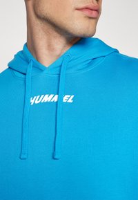 Felpa blu brillante con cappuccio e lacci, texture a coste e logo bianco "HUMANEL" sul petto. Presenta un design classico a pullover.