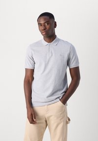 Grå bomulds polo skjorte med krave, korte ærmer og diskret logo broderi. Båret med lyse beige bukser med løs pasform.