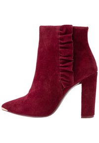 Stivale alla caviglia in suede bordeaux con punta appuntita, un accento arricciato sul lato e un tacco spesso. Finiture in tonalità oro sulla punta.