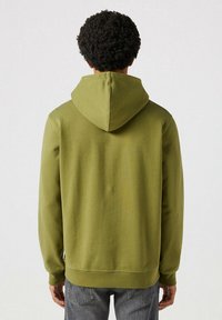 Sudadera verde oliva con un bolsillo canguro, puños acanalados y capucha con cordones. Tejido suave con una textura lisa. Vista trasera.