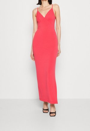 Woman wearing a fitted, sleeveless, floor-length coral dress with thin straps and black open-toe heeled sandals against a plain background.