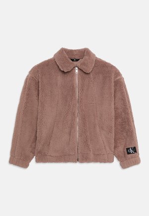 Veste zippée en peluche marron avec col et manches longues, avec un écusson logo noir sur le poignet gauche.