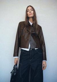 Chaqueta corta de cuero marrón con cierre de botón, combinada con una camisa blanca y pantalones acampanados de mezclilla oscura. Diseño sencillo, textura suave.