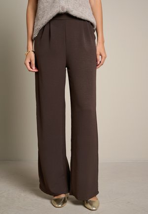 Vrouw draagt losse brede bruine broek met een beige gebreide top in de broek gestopt en gouden platte schoenen tegen een effen achtergrond.