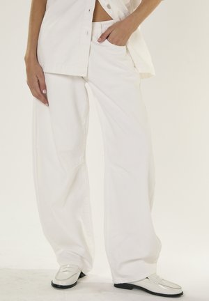 Kvinde iført løstsiddende hvide bukser og matchende hvid skjorte, stående med den ene hånd i lommen og hvide loafers på en ensfarvet baggrund.