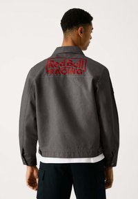 Graue Baumwolljacke mit einem auffälligen roten gestickten "Red Bull RACING" Logo auf dem Rücken. Klassischer Kragen und Knopfmanschetten.