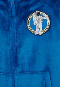 Zila vilnas zip-up kapucoja ir aprīkota ar apaļu uzlīmi ar astronauta ilustrāciju un tekstu "Kosmosa pētnieks misijā" kontrastējošā dizainā.