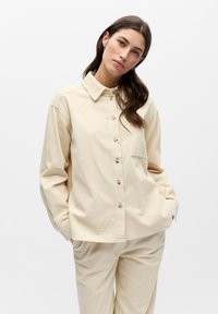 Chemise beige à boutons en tissu texturé, avec col, manches longues et une poche poitrine unique. Associée à un pantalon beige texturé.