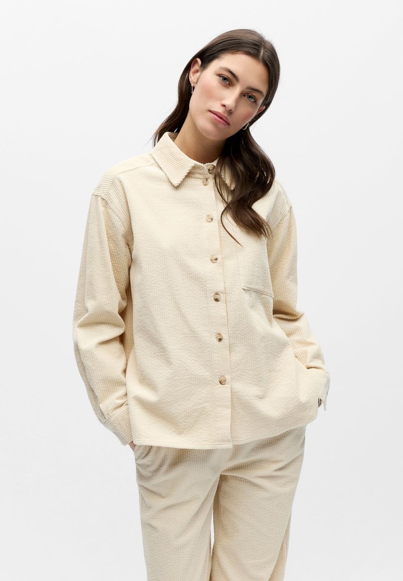 Chemise beige à boutons en tissu texturé, avec col, manches longues et une poche poitrine unique. Associée à un pantalon beige texturé.