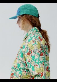 Jeune personne aux cheveux roux attachés en queue de cheval, portant une casquette verte et une veste colorée à motif floral, face à gauche sur un fond clair uni.