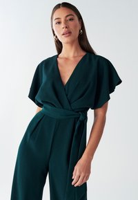 Tumši zaļa aplieciena jumpsuīts ar īsām plandošām piedurknēm, dziļu V-veida izgriezumu un sasienamu vidukli. Gluda auduma ar brīvu piegriezumu un platiem bikšu galiem.