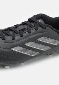 Černý fotbalový kopač s texturovaným svrchním dílem, obsahující tři stříbrné reflexní pruhy a černé tkaničky. Hroty kopaček jsou bílé.