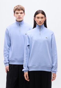 Ανοιχτό γαλάζιο φλις φούτερ με μισή φερμουάρ και ρ ιμπ υφή, με μικρό πράσινο λογότυπο στο στήθος και άνετη εφαρμογή, συνδυασμένο με σκούρο παντελόνι.