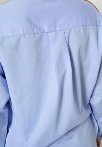 Svetlomodrá košeľa s dlhými rukávmi, ktorá má štruktúrovaný chrbát s vertikálnymi záhybmi a hladkou tkanou textúrou. Klasický golier.