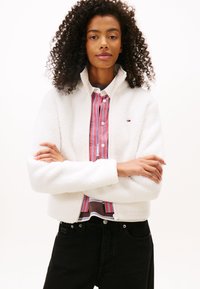 White fleece jacket with a high collar, cropped design, and ribbed cuffs, paired with a striped shirt. Black jeans complete the outfit.