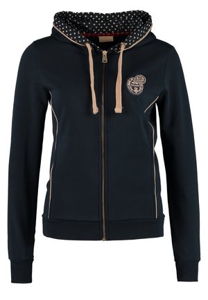 Frau trägt beige Hose, blau gemustertes Oberteil, schwarzen Reißverschluss-Hoodie mit gepunktetem Innenfutter und glänzende schwarze Stiefeletten, steht mit einer Hand in der Tasche.