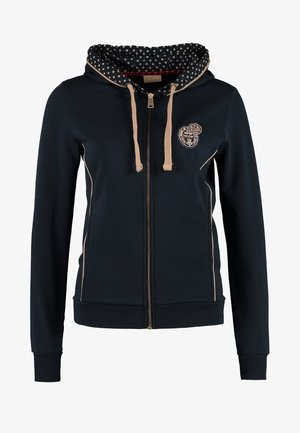 Femme portant un pantalon beige, un haut bleu à motifs, un sweat à capuche noir zippé avec doublure à pois, et des bottines noires brillantes, debout avec une main dans la poche.