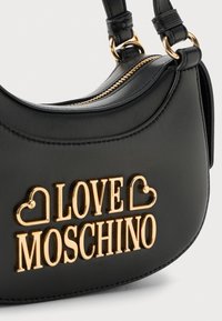 Bolsa de mão em pele preta com formato curvado, logótipo dourado "LOVE MOSCHINO", fecho com zíper e alça curta com detalhes em metal dourado.
