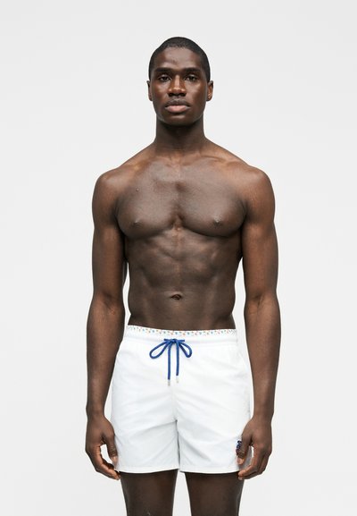 Μυώδης άνδρας με σκούρο δέρμα που στέκεται με το πρόσωπο προς τα εμπρός, φοράει λευκά μαγιό με μπλε κορδόνι, μπροστά σε απλό φωτεινό φόντο.