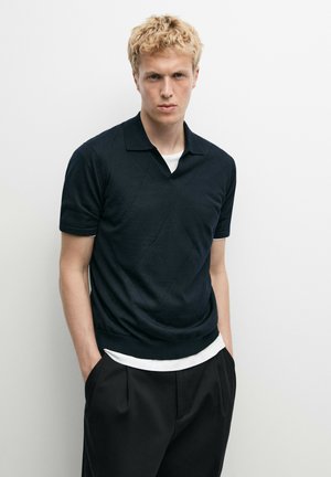Giovane uomo con capelli biondi corti che indossa un maglione nero a maniche corte sopra una camicia bianca e pantaloni neri, in piedi davanti a uno sfondo bianco.