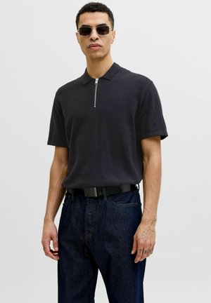 Mand iført sort kortærmet polo med lynlås foran, mørke jeans, sort bælte, solbriller og synlige tatoveringer på højre hånd og fingre.