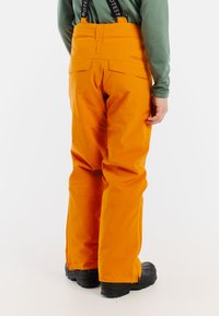 Oranje waterdichte skibroek met verstelbare bretels, met een recht model en verstevigde enkels. Zwarte schoenen zichtbaar.