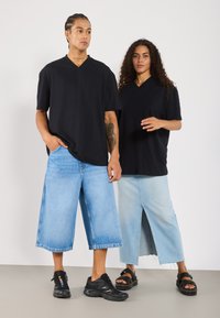 Sorte oversized v-hals t-skjorter på to modeller. En har på seg blå vide bukser, mens den andre har på seg et lyseblått skjørt med splitt foran, begge med svarte sko.