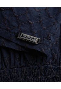 Námornická prošívaná látka s diamantovým vzorem, se stříbrnou značkovou destičkou "Superdry" a elastickým pasem se shromážděnou texturou.