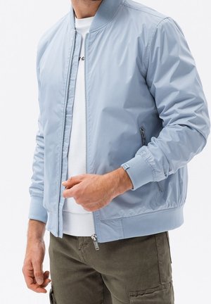 Mężczyzna w jasnoniebieskiej kurtce bomberce na białej koszuli i oliwkowych spodniach typu cargo, z częściowo rozpiętym zamkiem i rozluźnionymi rękami.