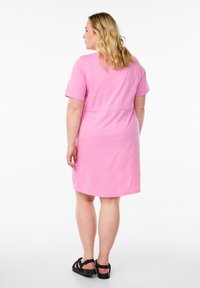 Robe en coton rose à manches courtes, avec une coupe décontractée et une taille cintrée. Associée à des sandales noires, vue de dos.