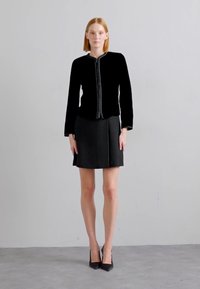 Chaqueta de terciopelo negro con un diseño ajustado y cremallera en contraste, combinada con una falda mini negra. Tacones altos completan el conjunto.