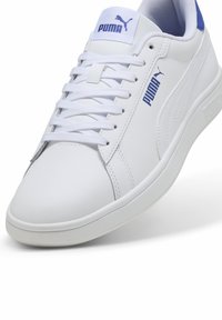 Sneaker in pelle sintetica bianca con accenti blu, punta arrotondata e suola piatta. Le caratteristiche includono lacci piatti e il logo Puma sulla linguetta e sul lato.