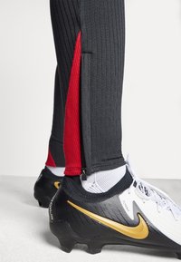 Detailní záběr na černé a červené sportovní kalhoty se bočním zipem nad bílými ponožkami a černo-bílými fotbalovými kopačkami se zlatým logem Nike.