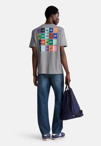 Šedé bavlněné tričko s barevnou grafikou ryby na zadní straně, spárované s modrými džíny a tmavě modrou taškou na tkaničku.