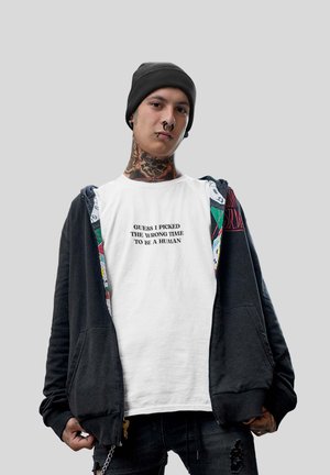 Sort zip-up hættejakke over en hvid t-shirt med tekst, parret med mørke jeans. Inkluderer en beanie og synlige tatoveringer.