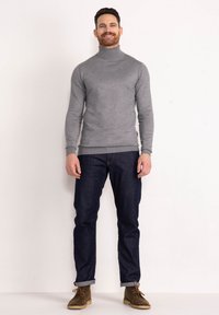 Grauer Rollkragenpullover mit langen Ärmeln, kombiniert mit dunkelblauen Jeans und braunen Stiefeln, vor einem weißen Hintergrund stehend.