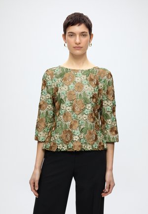 Ung kvinde med kort mørkt hår iført gyldne øreringe og en gennemsigtig bluse med blomsterbroderi i beige, grønne og brune nuancer.